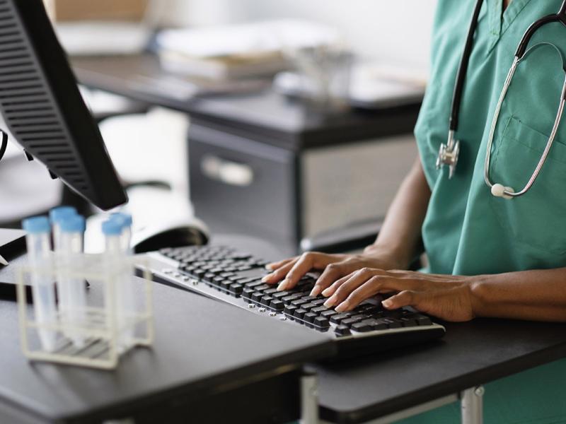  Health care professional wearing scrubs and a stethoscope typing on a computer desktop.