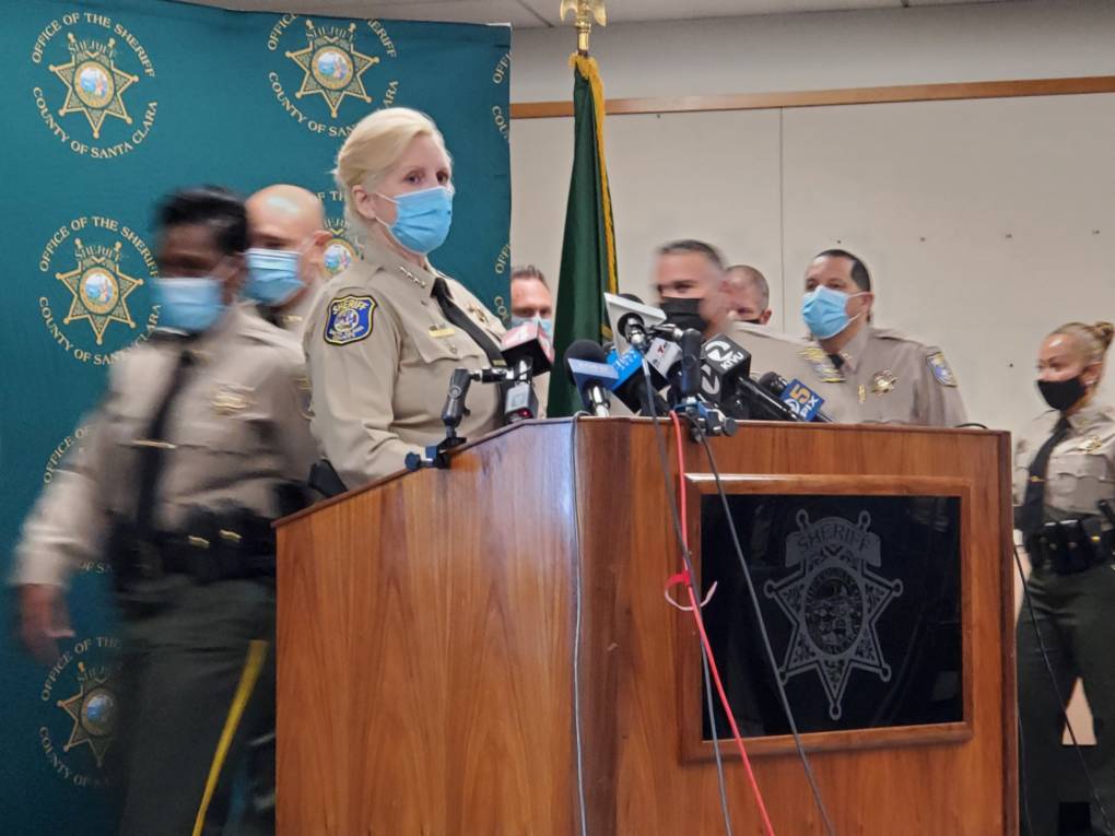 The female sheriff, in a light brown uniform and wearing a mask, flanked by many staff members in the same outfit, stands at a podium inside.