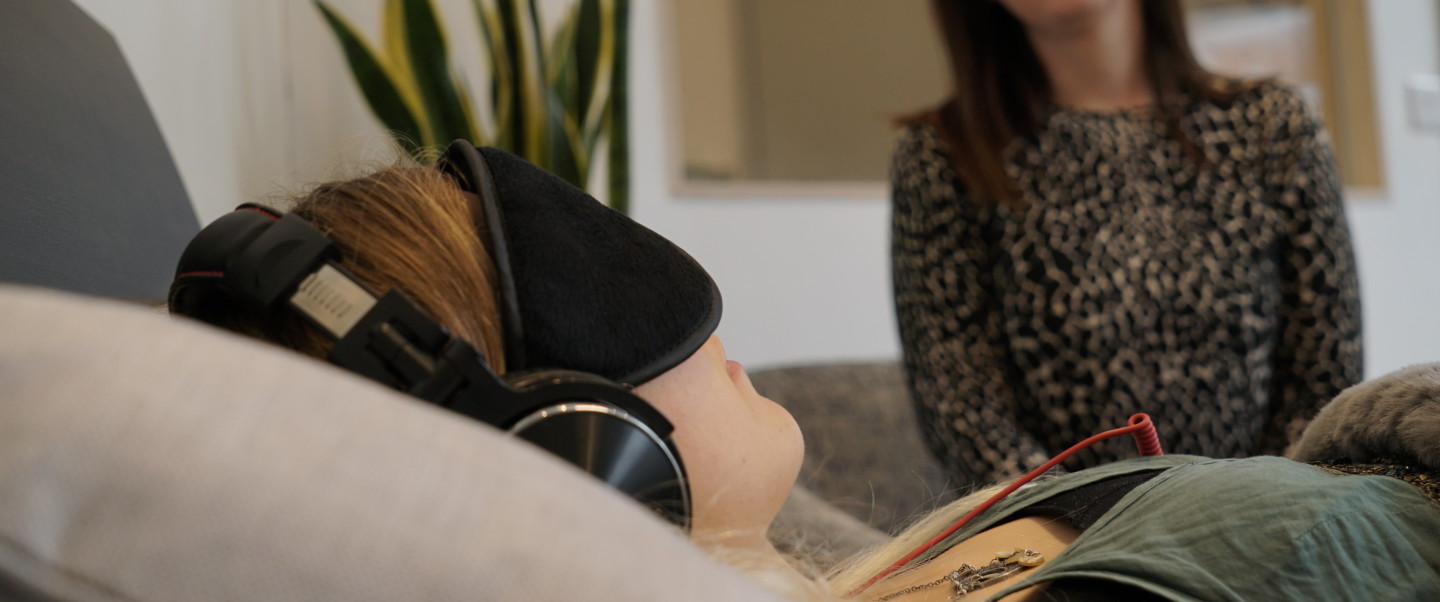 Light-skinned woman lying on sofa wearing black eye mask and headphones. A light-skinned woman sitting nearby overlooks her. Therapy setting.