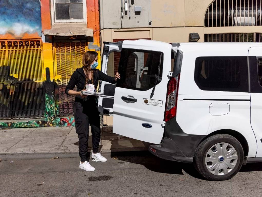 A woman closes the back doors of a van after taking out some equipment. The van is parked on the street in front of a row of houses.