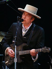 Bob Dylan plays a guitar and sings into a microphone.