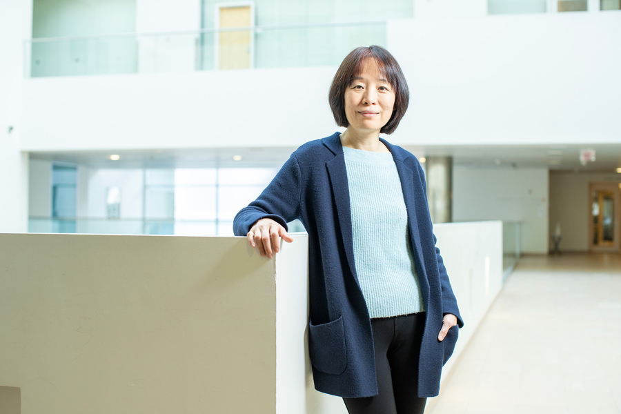 Fan Wang is wearing a blue cardigan, has one hand in her pocket and the other resting on a low wall inside the Brain and Cognitive building. 