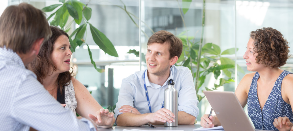 A group of people sat around a table having a discussion. On the table is a laptop