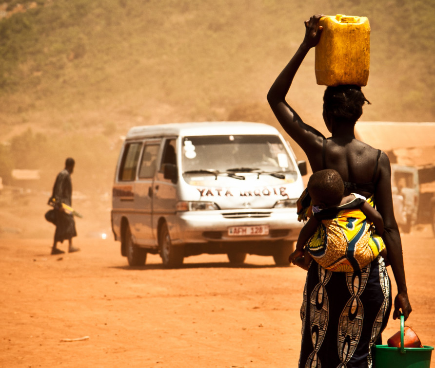 Photo of woman in Africa carrying water and walking towards a bus that is heading towards us the viewers.