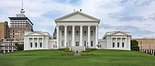 All white Neoclassical building with pediment and six columns rises on a grassy hill with a large American elm tree in the left foreground. Two boxier, but similarly styled wings are attached at the building's rear.