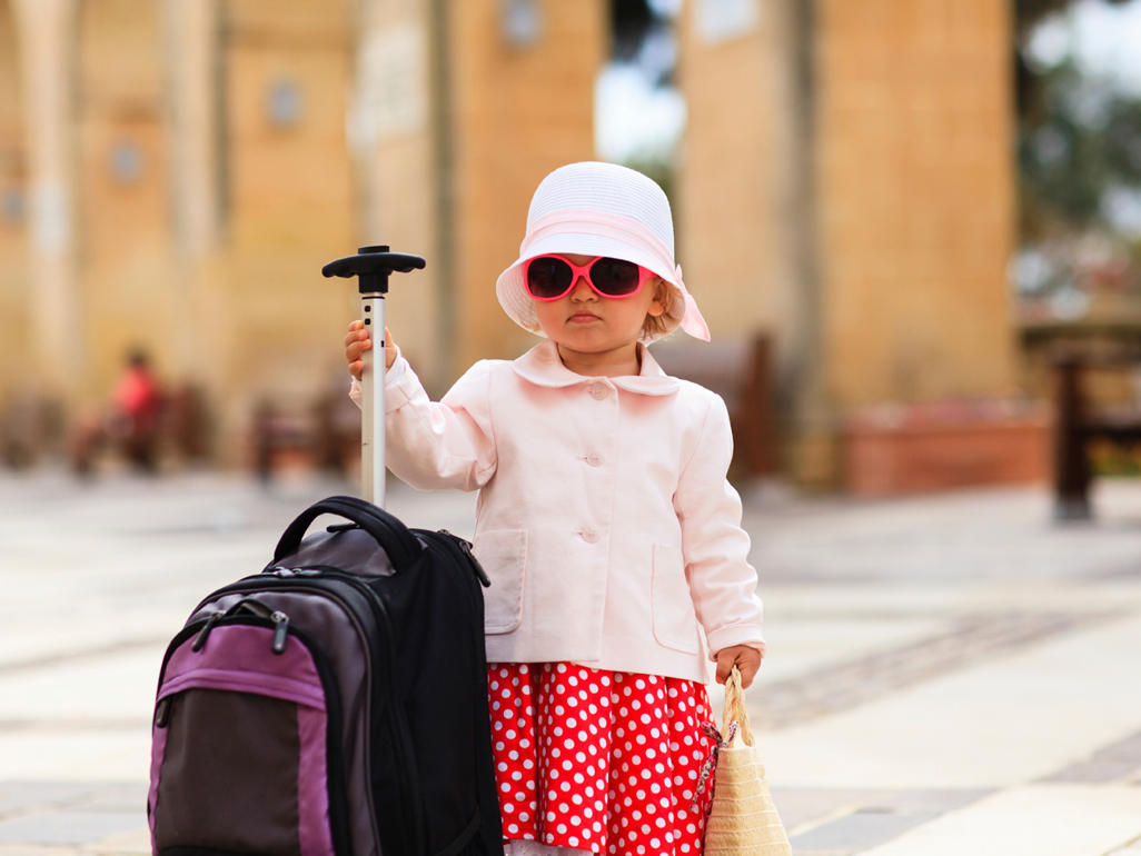 toddler wearing hat and sunglasses and standing beside a suitcase