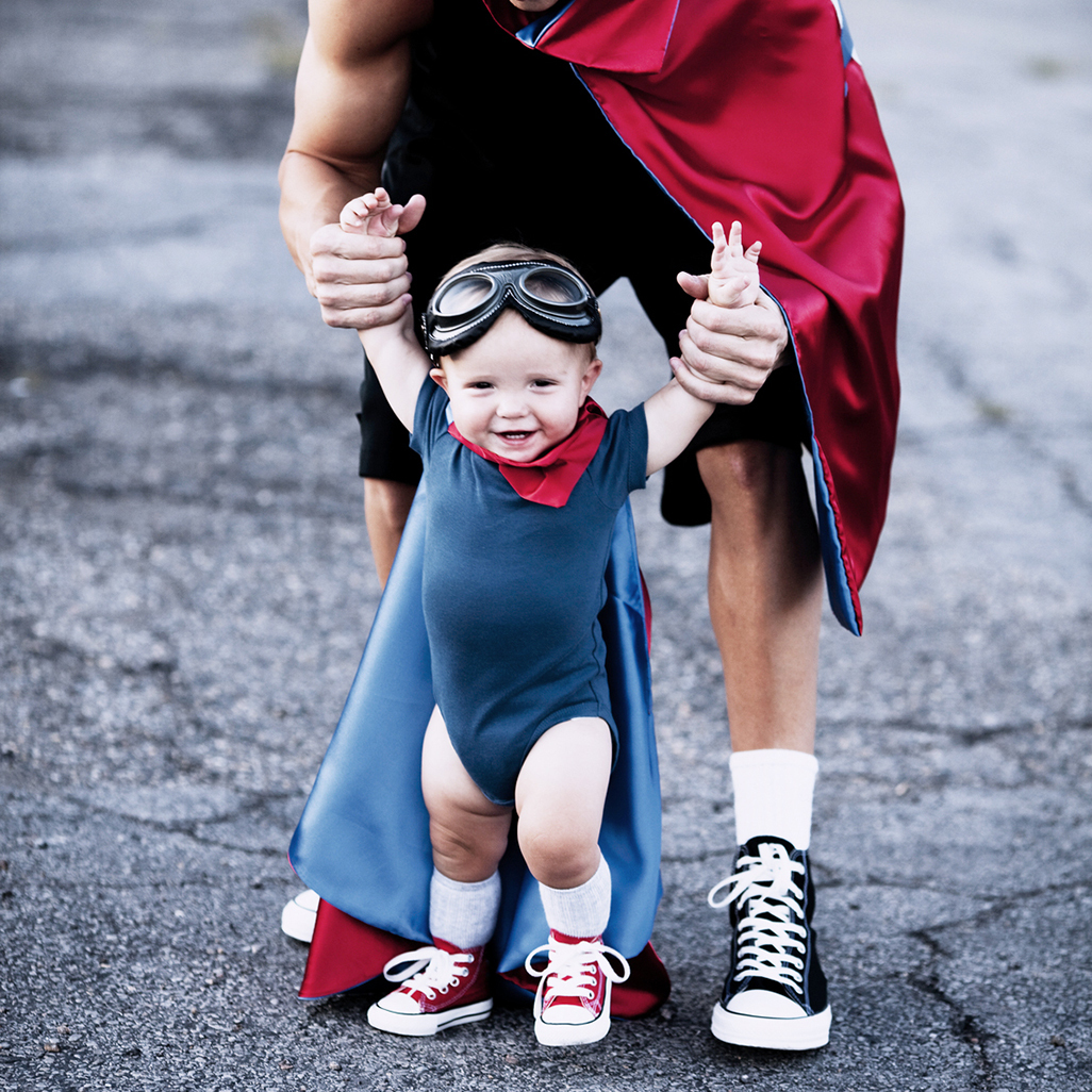 baby dressed in super hero costume with it's arms held up by adult