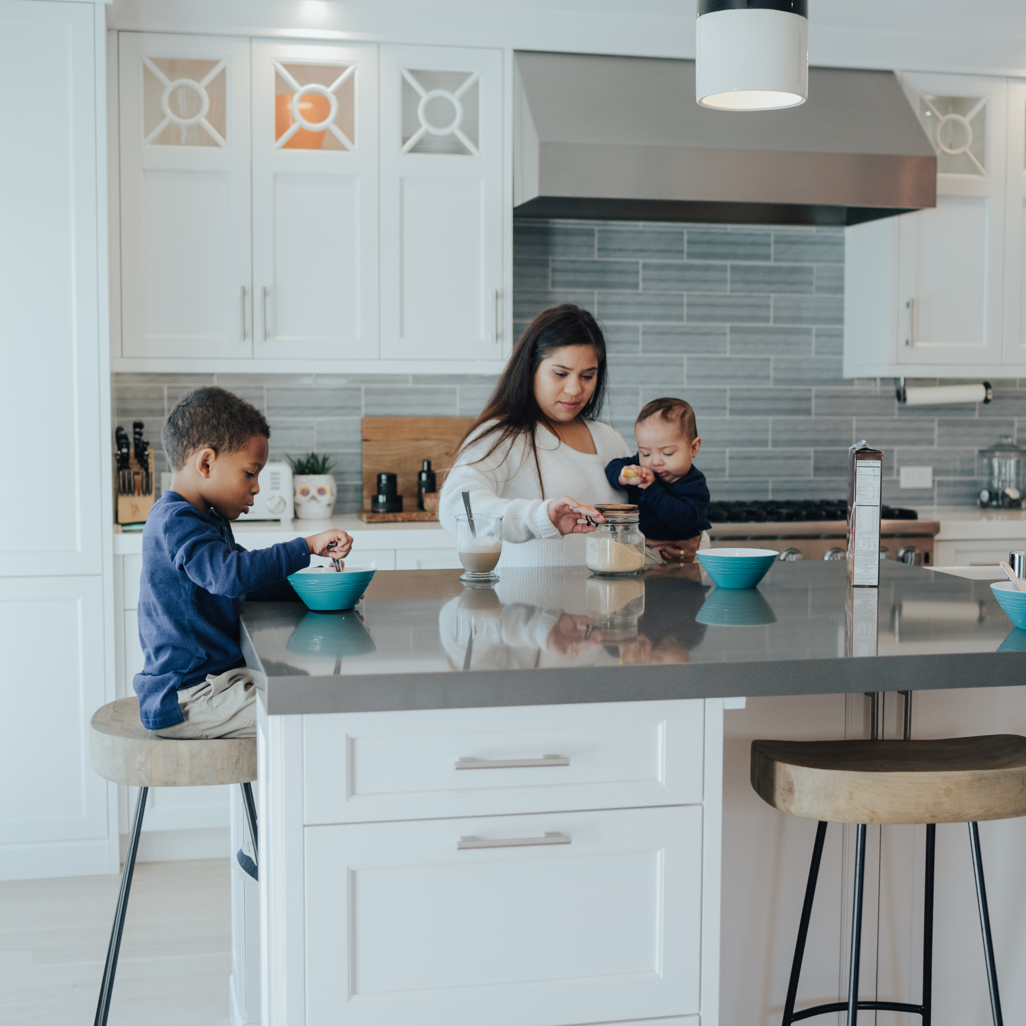 A mom standing at her kitchen counter, holding her baby while a toddler eats cereal.