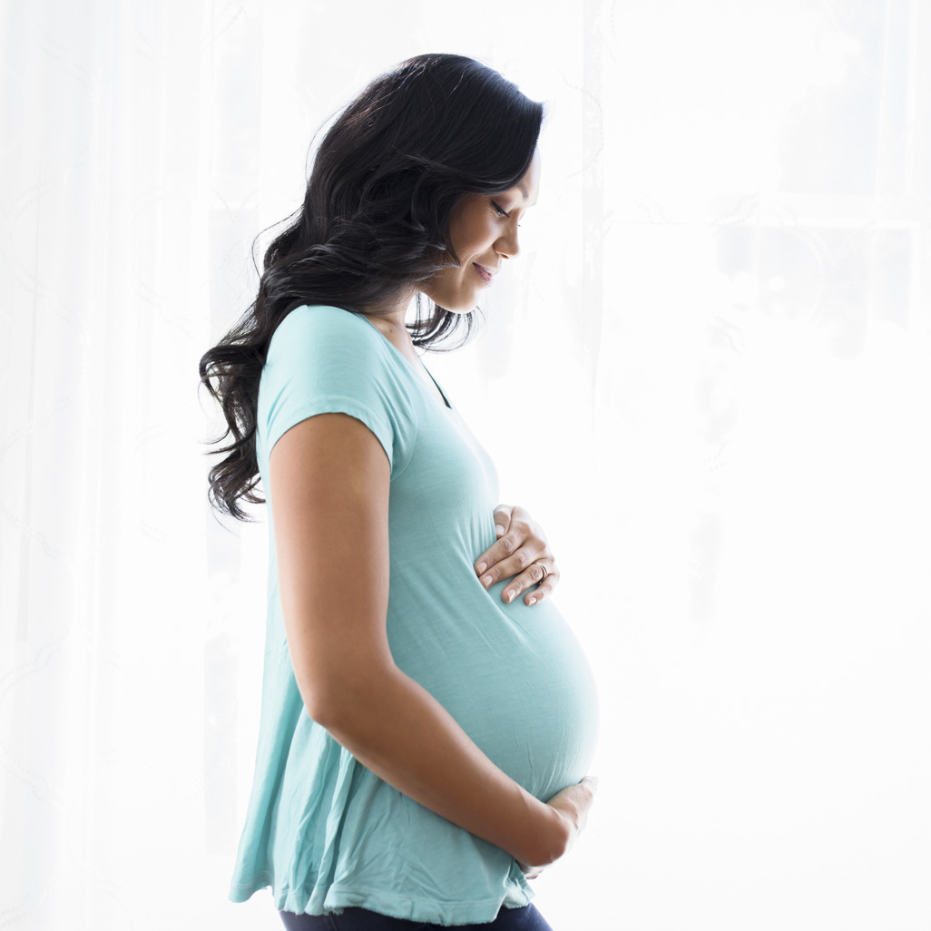 A pregnant woman standing in front of a window, cradling her belly.