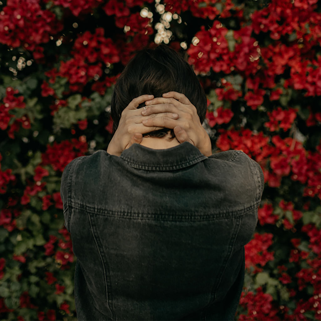 The back of a woman, who's holding her head in her hands and facing a wall of flowers.