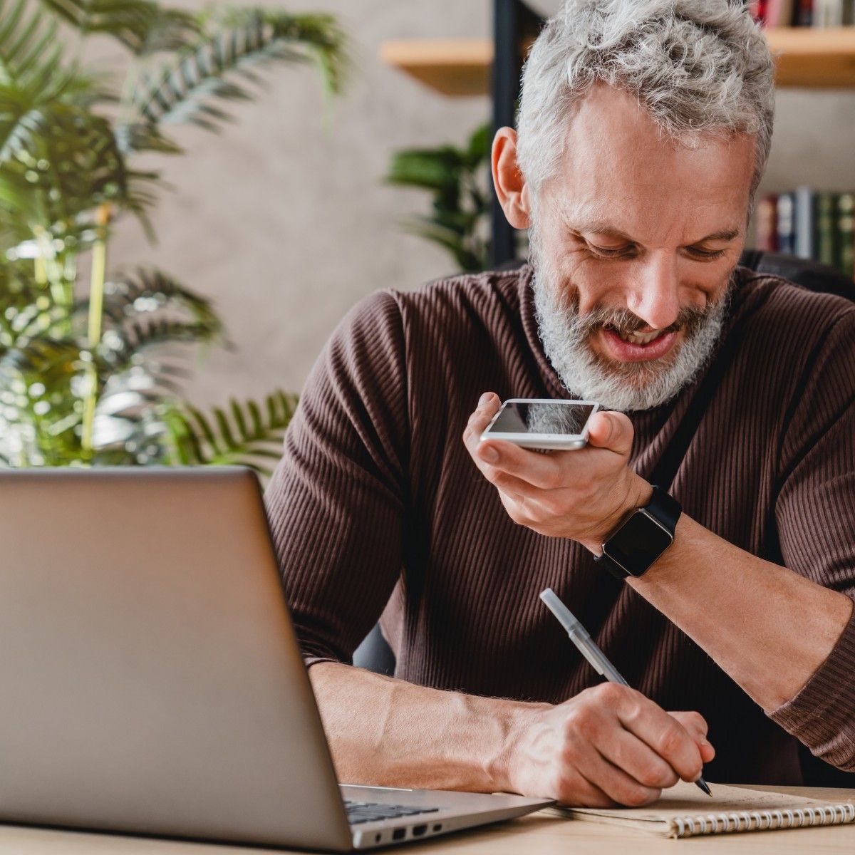 A senior man writes on a notepad while talking on speakerphone and sitting in front of a laptop