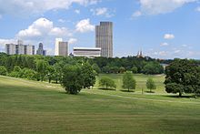 A green space with trees and rolling lawns is flanked by tall, modern-style buildings in the background on a sunny day.