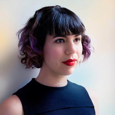 Caroline Sinders. Research Fellow, Harvard Kennedy School. Wearing a black shirt, in front of a light colored wall.