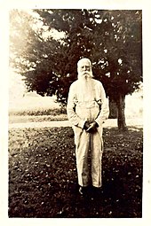 A black and white image (c. 1900s) of an elderly man with white hair and a long beard dressed in a long-sleeved and suspenders standing in front of a tree.