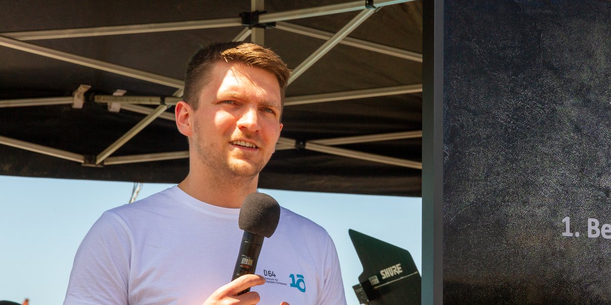 Foto von Erik Tuchtfeld in weißem Tshirt auf der re:publica 2022. Hinter ihm ist ein schwarzes Zelt erkennbar. Ein Monitor ist rechts aufgebaut, auf dem eine Präsentation läuft.