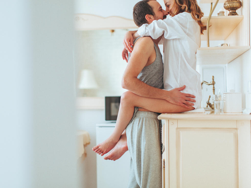 A man with his arms around the waist of a woman sitting on a kitchen counter. 