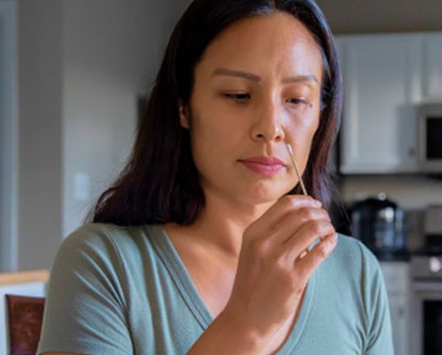 Person sitting in a chair in a kitchen swabbing their nose for an at-home COVID-19 test.