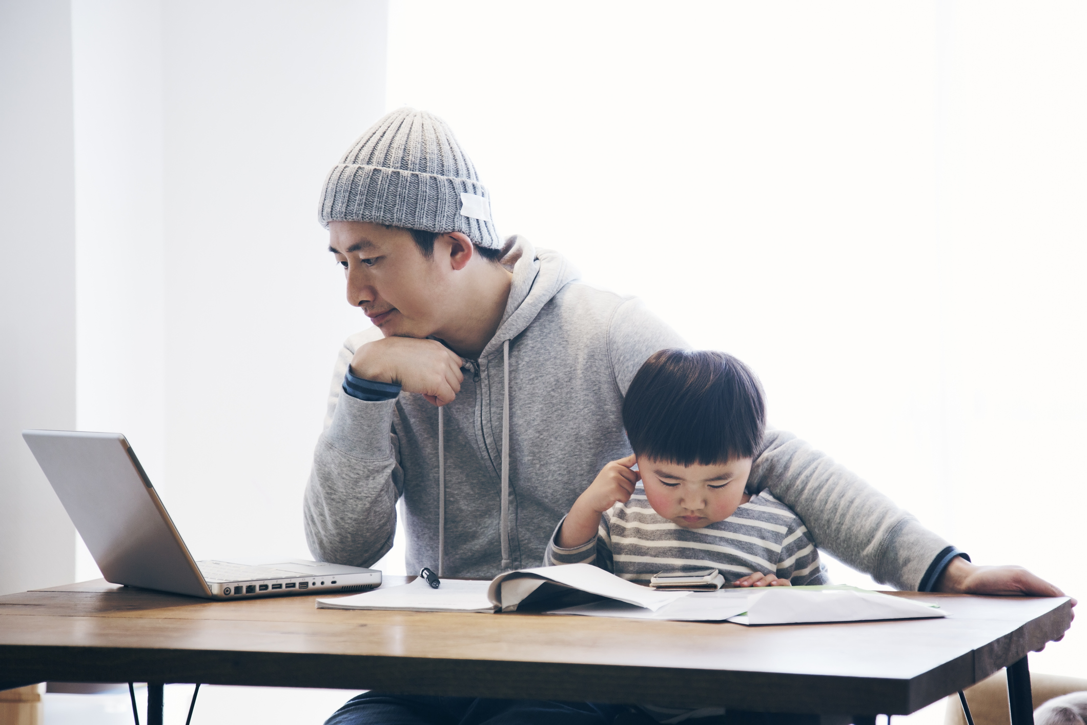 Japanese man in casual clothes using a laptop and his son using a smartphone on the desk. He's working and doing childcare at home.