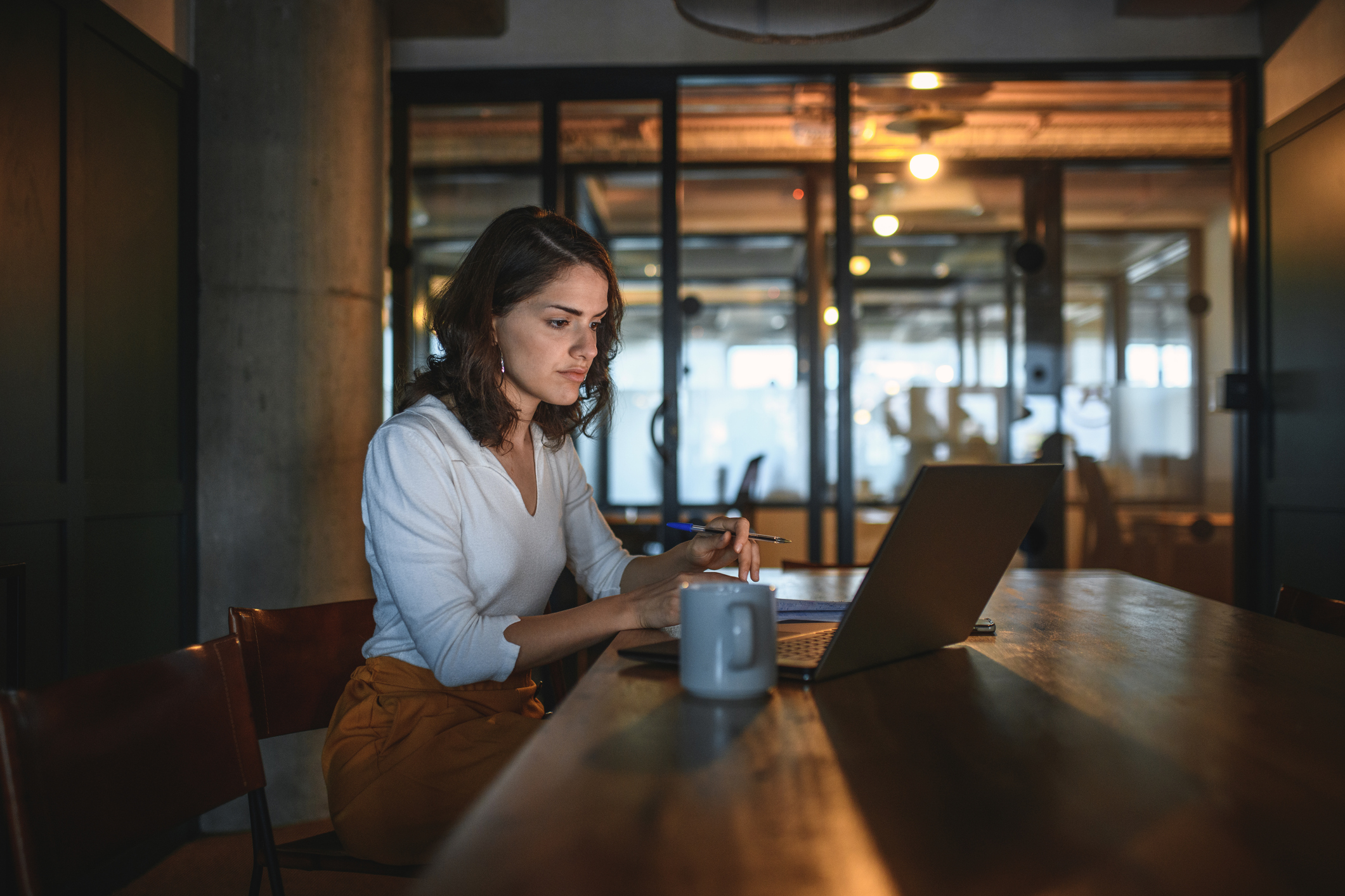 Brunette businesswoman in late 20s sitting alone with coffee and laptop on conference table in dimly lit board room.