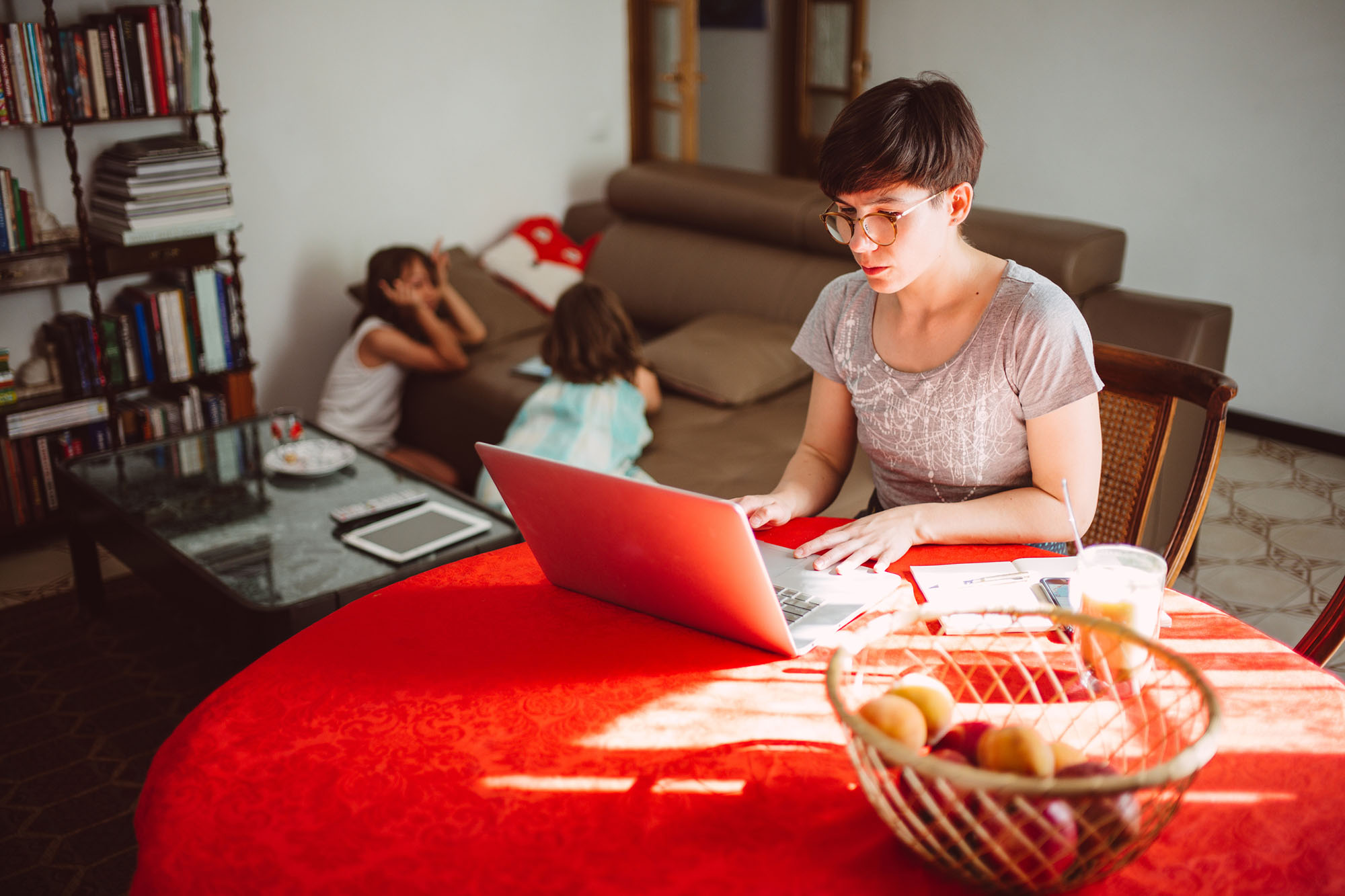 Mother working from home at kitchen table with children playing in the background on couch