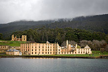 A calm body of water is in the foreground. The shoreline is about 200 metres away. To the left, close to the shore, are three tall gum trees; behind them on an incline are ruins, including walls and watchtowers of light-coloured stone and brick, what appear to be the foundations of walls, and grassed areas. To the right lie the outer walls of a large rectangular four-storey building dotted with regularly spaced windows. Forested land rises gently to a peak several kilometres back from the shore.