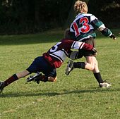 A child running away from camera in green and black hooped rugby jersey is being tackled around the hips and legs by another child in opposition kit.