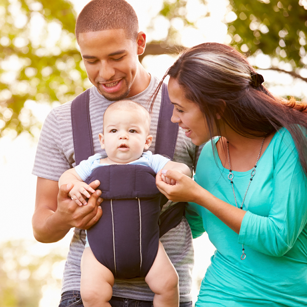 parents walking outside, father carrying their baby in a carrier, while mother is holding baby's hand