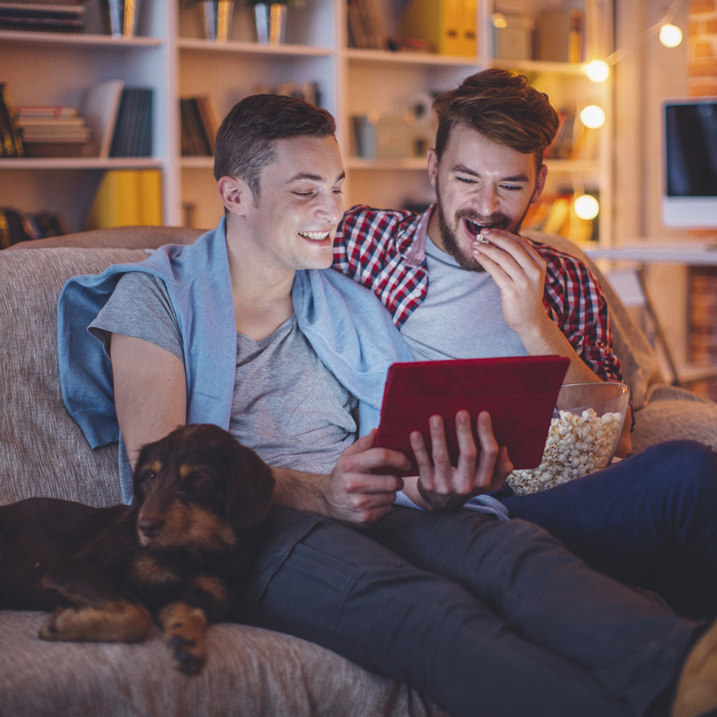 two adult men sitting on a couch with their dog while watching a tablet device and eating popcorn