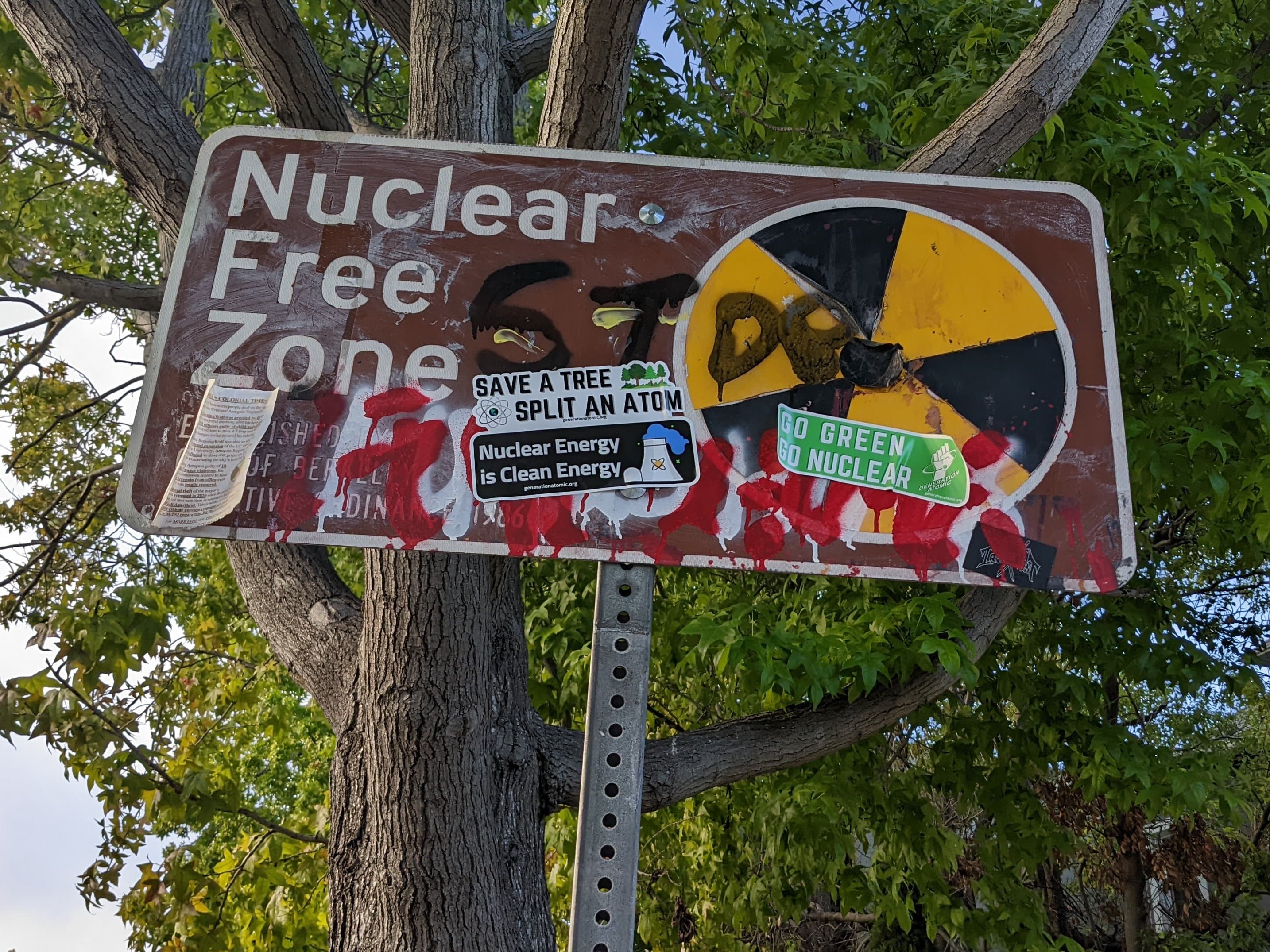 A street sign at the edge of the City of Berkeley, California, that says "Nuclear Free Zone." The sign has been heavily defaced including several pronuclear stickers with slogans such as "Go Green, Go Nuclear" and "Save a tree, split an atom" and "Nuclear energy is clean energy."