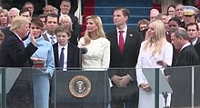 Trump, surrounded by dignitaries, raises his right hand and places his left hand on the Bible as he takes the oath of office. John Roberts stands across from him administering it.