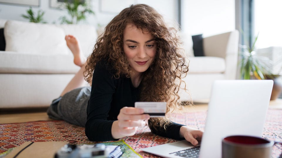 Woman lying on floor with credit card in hand looking at laptop. 