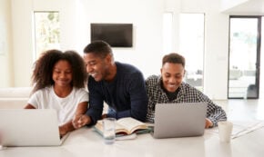 The photo shows three people, presumably (left to right) teen daughter, father, and teen son. The three are working on the teens' laptop computers on their kitchen counter at home. The room is mostly white with bright sunshine.
