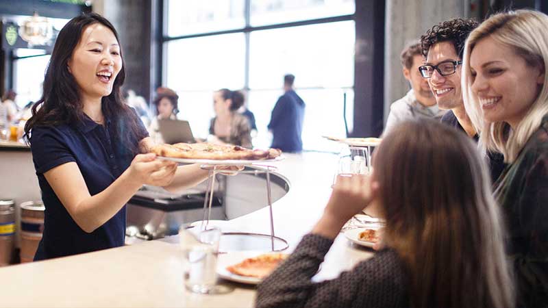 Woman serving pizza to a group of people at a bar.