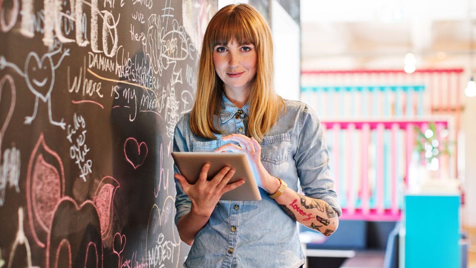 Female small business with tattoos leaning against chalkboard with tablet in hand.