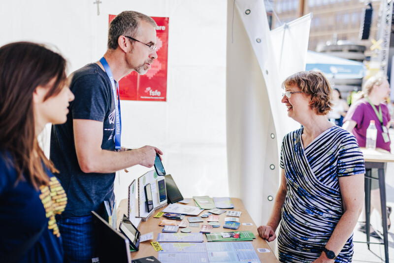 Person pointing to one out of four old phone devices demonstrated in a booth, two people listening.