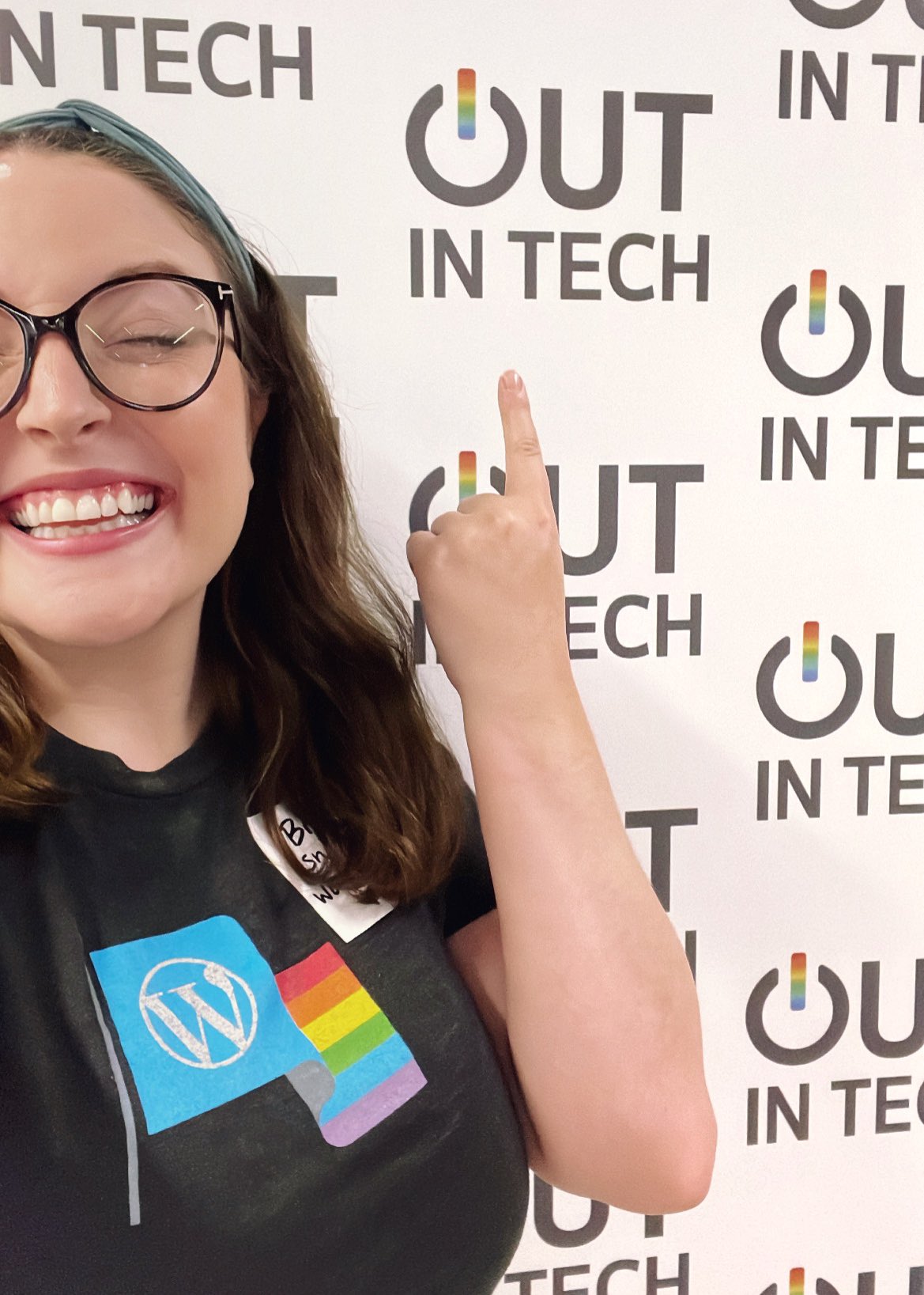 Bre in front of the Out in Tech backdrop. Wearing a gray shirt with a pride flag WordPress shirt.