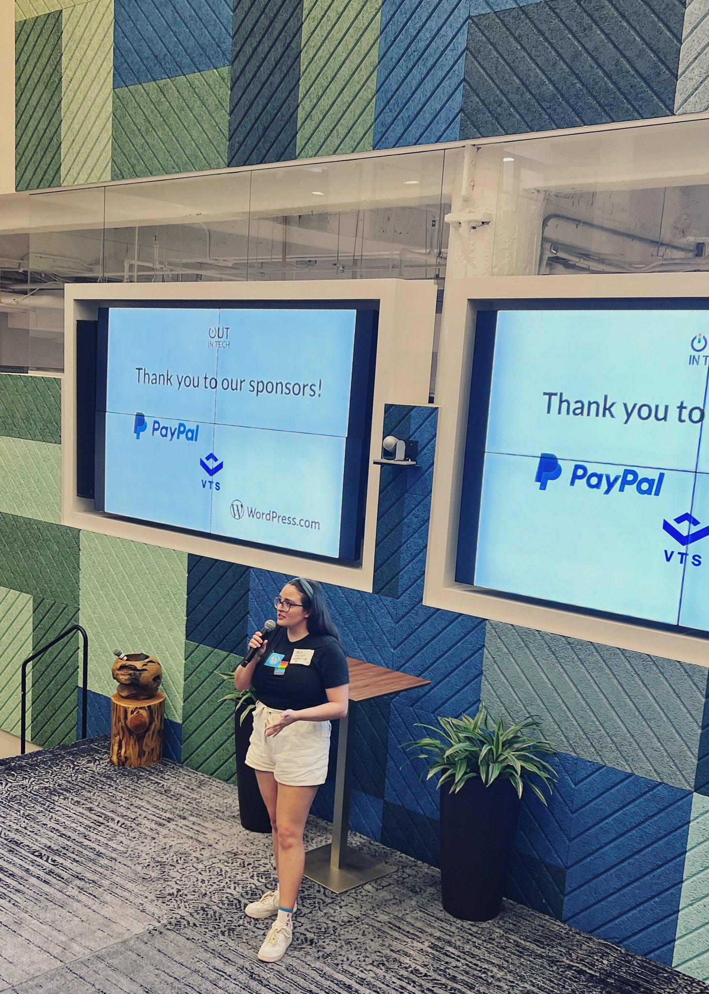 Bre speaking on a stage. She is wearing white shorts, a pride flag WordPress shirt, a blue headband, and white shoes. On the screen behind, the text reads: Thank you to our sponsors! PayPal, VTS, WordPress.com