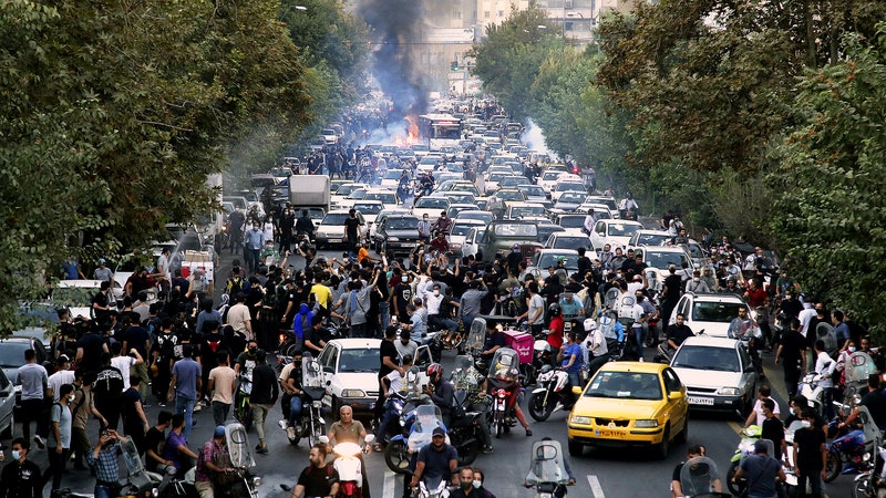 A city street in Iran filled with protestors and cars. Fire and smoke can be seen in the distance.