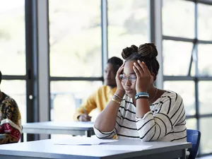 Worried young female student sitting with head in hands at desk during exam in community college classroom