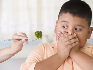 young boy covering mouth as hand tries to feed him broccoli on fork