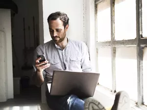 a man looking at his phone and computer on a windowsill