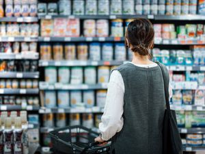 Rear view of young Asian mother with a shopping cart grocery shopping for baby products in a supermarket. She is standing in front of the baby product aisle and have no idea which product to choose from