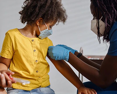 Young child holding parent’s hand, wearing a mask looking down at their arm as doctor applies a bandage.