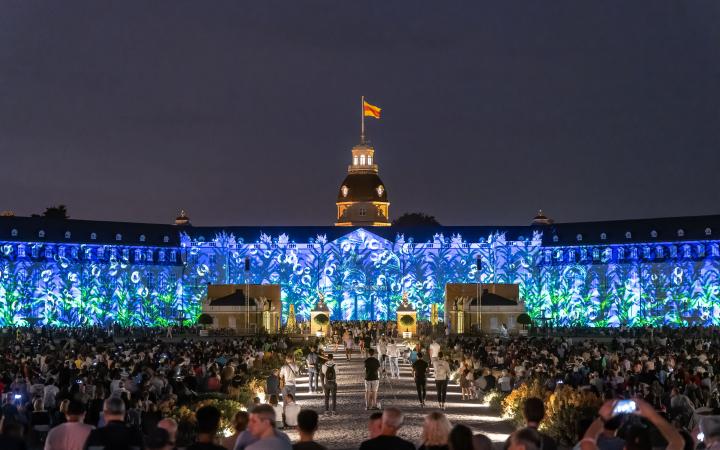 Das Karlsruher Schloss erleuchtet in einem blau-grünen Pflanzenmuster. Zu sehen ist auch die vielen Menschen, die im Publikum sitzen.