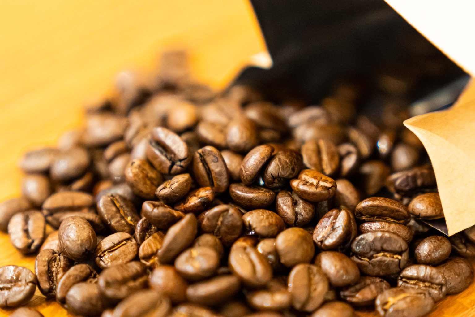 Close-up shot of coffee beans piled outside a bag on a yellow table.