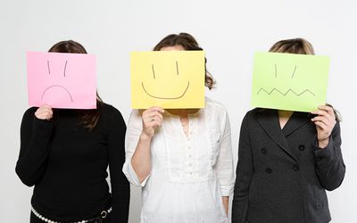 Three women holding papers over their faces with different expressions drawn on