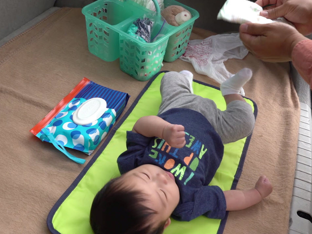 Baby is laying on a changing mat in the open boot of a car, while dad is holding a nappy