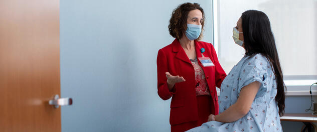 A female Mayo Clinic doctor talking to a seated female patient in a patient gown, both wearing masks.