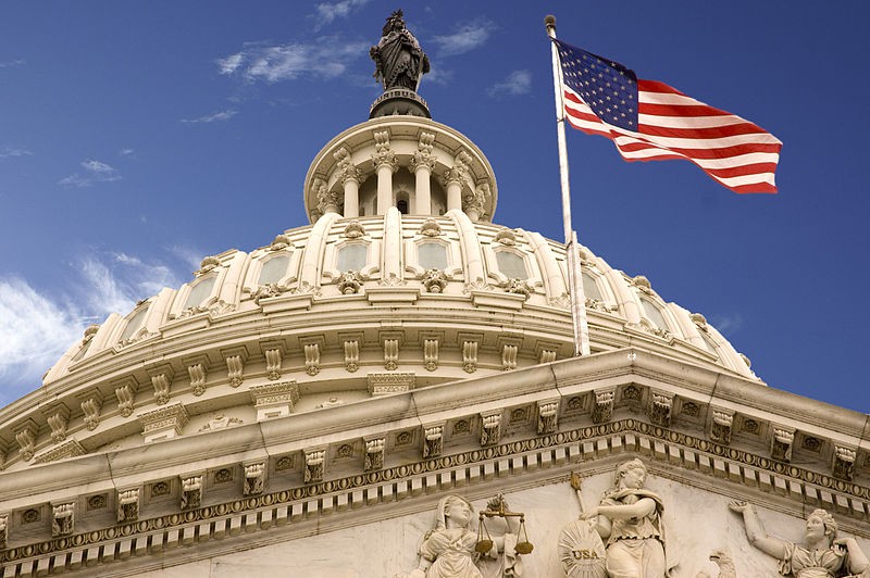 Image of the United States Capitol Building Dome, from a bottom-up perspective. Dome is white against a blue sky, with a big American flag waving in the wind.