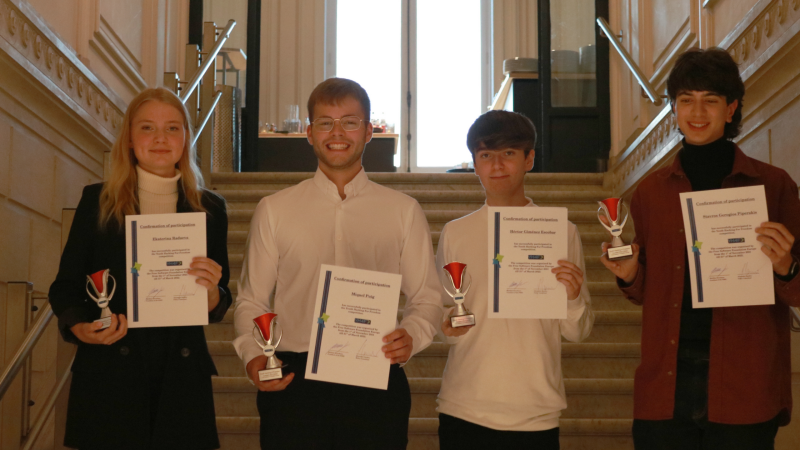 One young woman and three young men holding certificates and trophies, standing in front of stairs in a neoclassical building